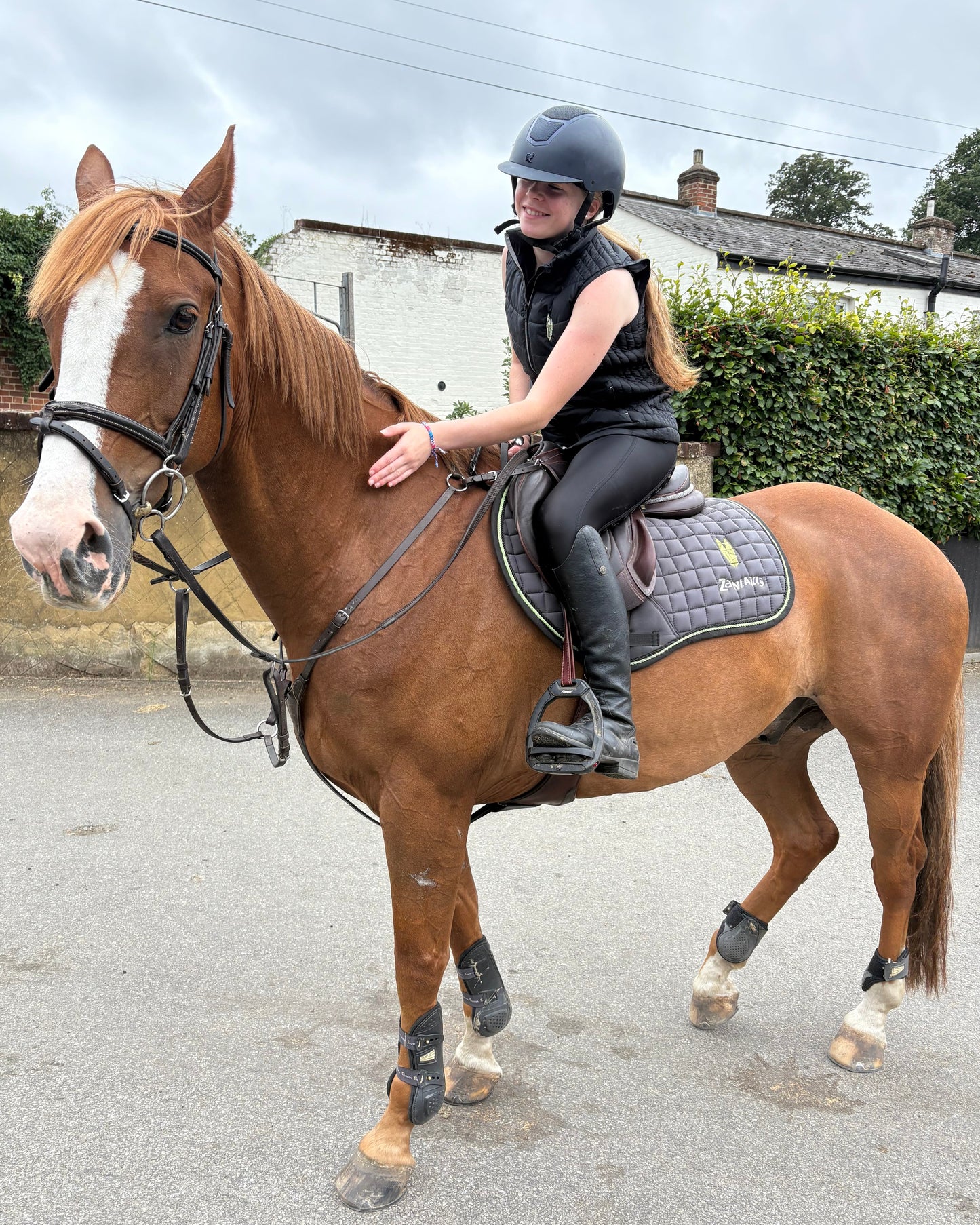 Woman riding a chestnut horse wearing a black equestrian gilet bodywarmer on a road with a house and trees in the background