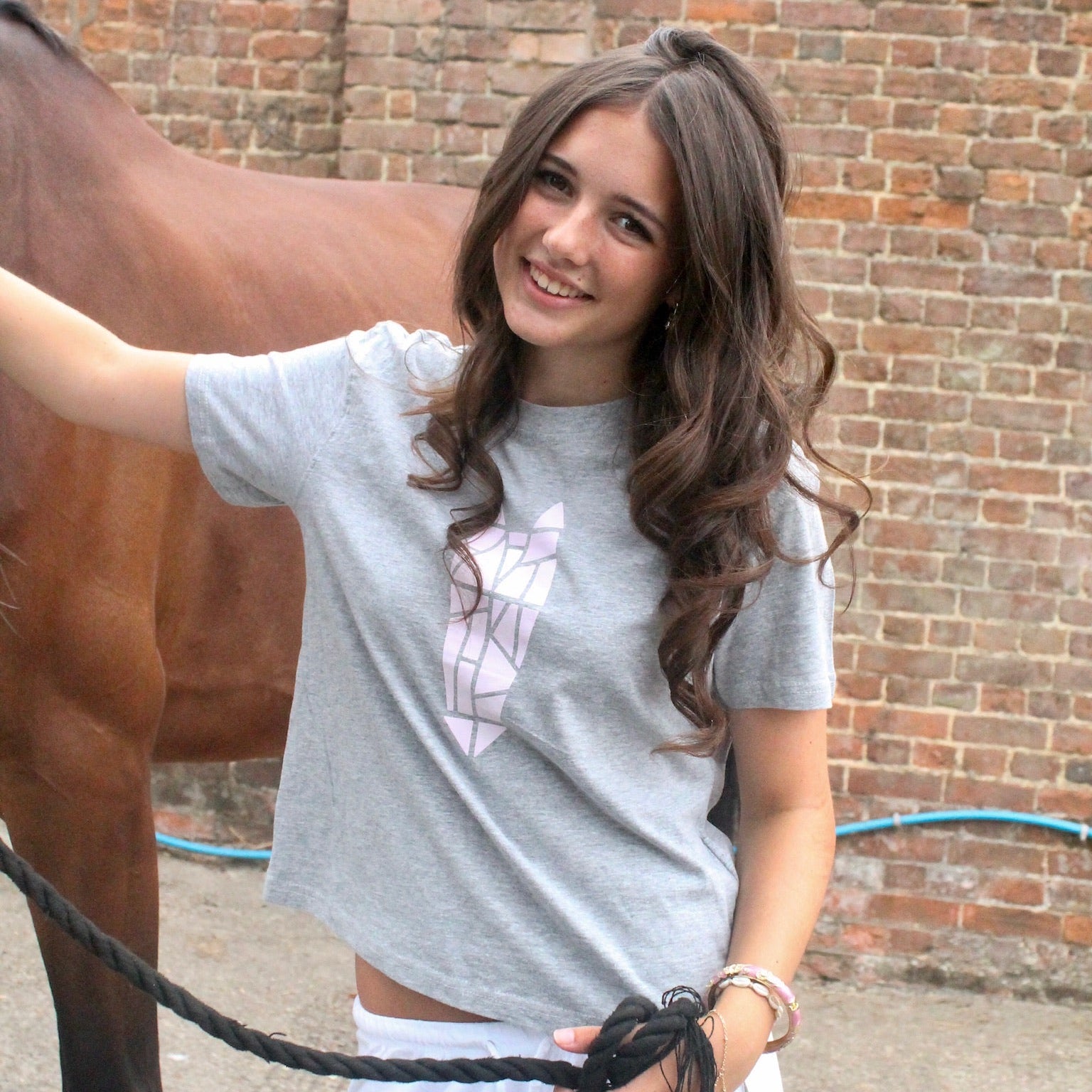 Woman holding a horse wearing a grey tshirt with a purple geometric horse head icon by a brick wall