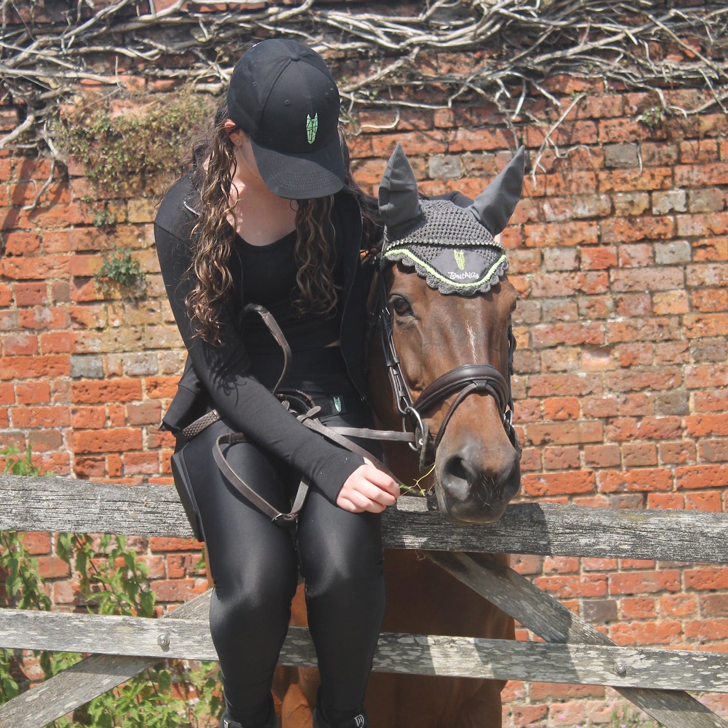 Teenage girl sitting on a fence with a horse wears an equestrian baseball cap with a distinctive silver green logo