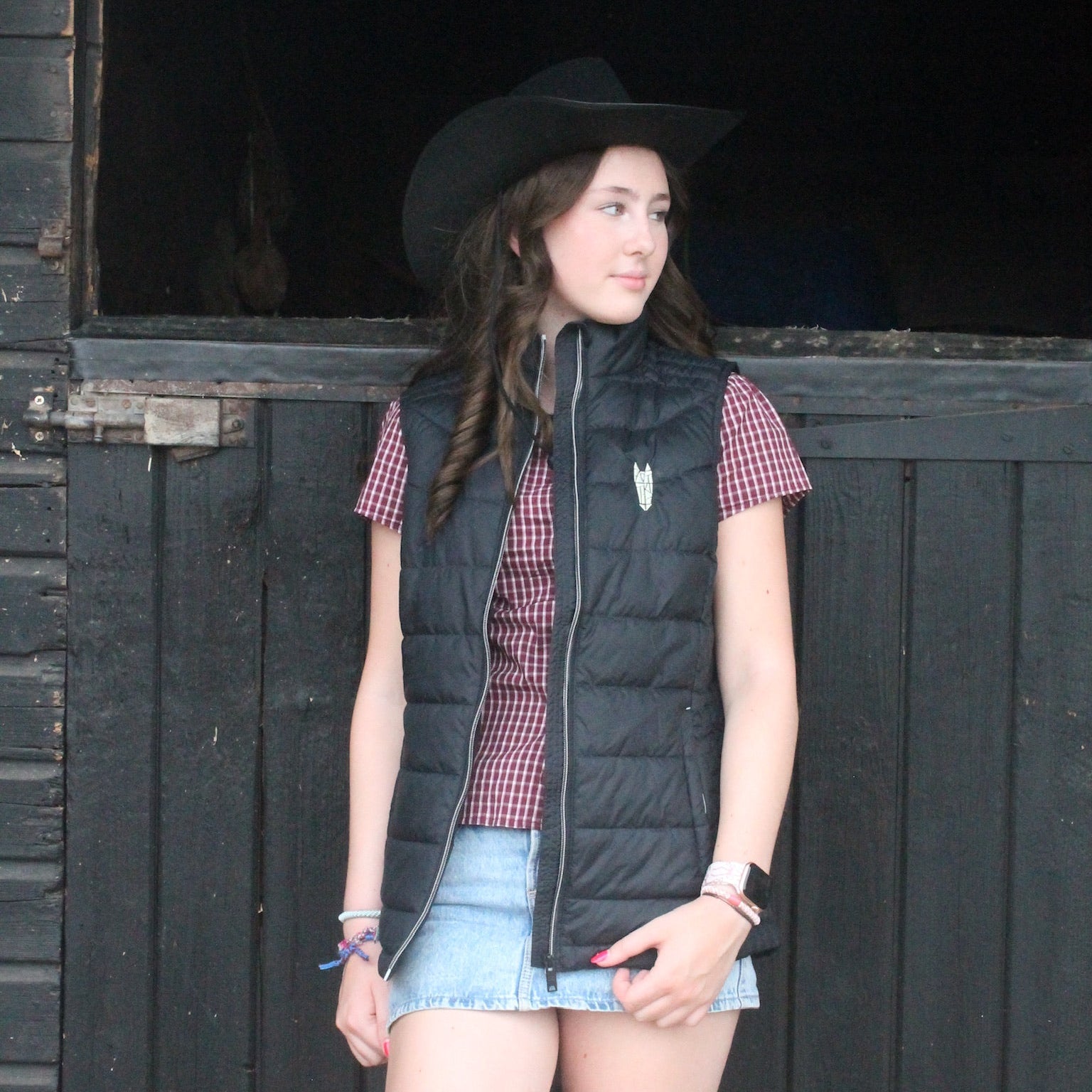 Woman wearing a black equestrian bodywarmer, plaid shirt, and denim shorts standing in front of a dark wooden stable door.