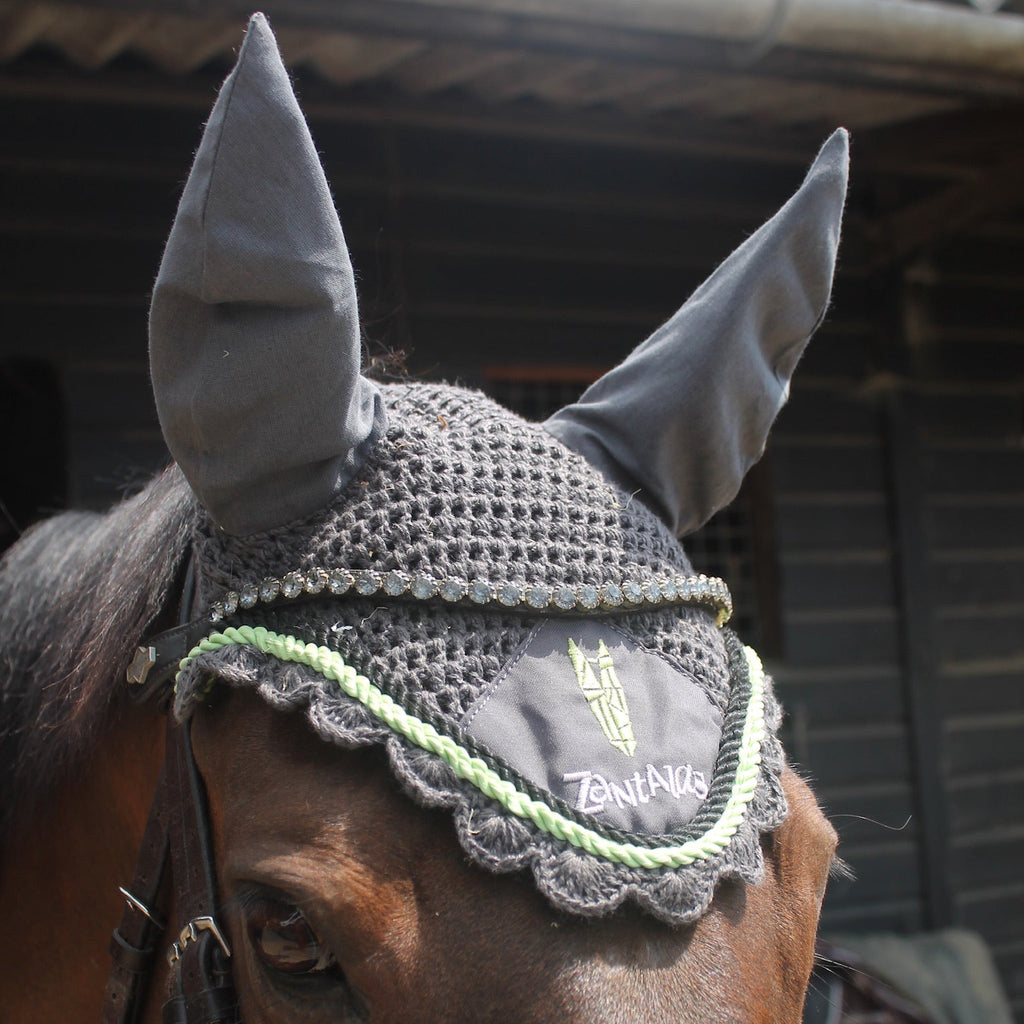 Horse wearing a gray crochet fly hood with green accents in front of a stable.