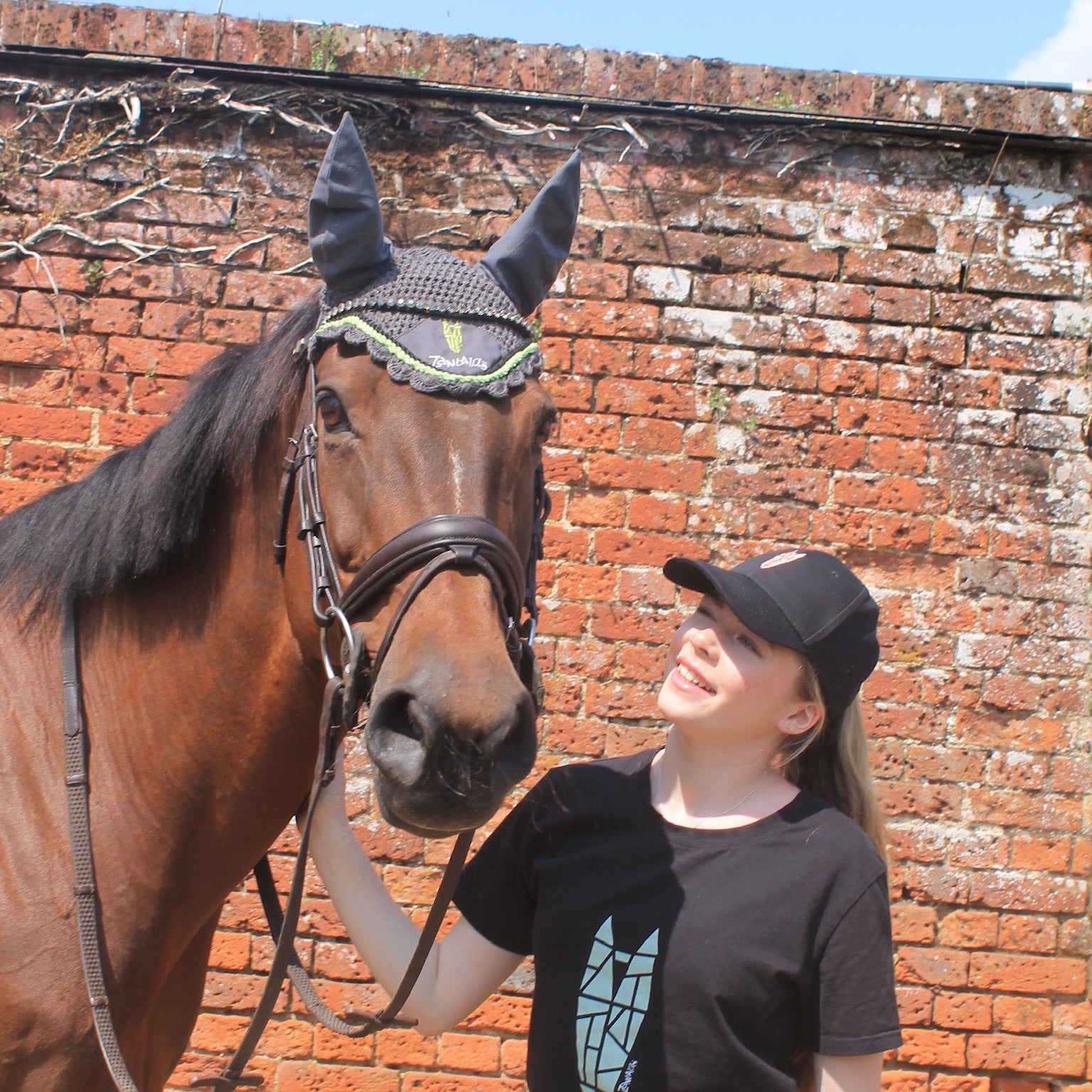 Woman standing next to a horse with a bridle against a brick wall.