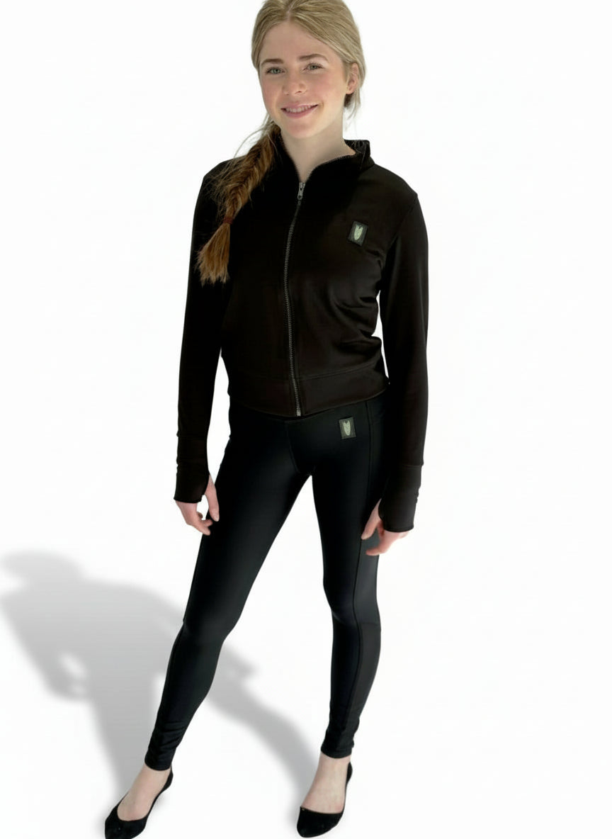 Full length image of a young woman wearing a black colour coordinated equestrian outfit with a light black jacket/cover up top and riding leggings with a mesh panel