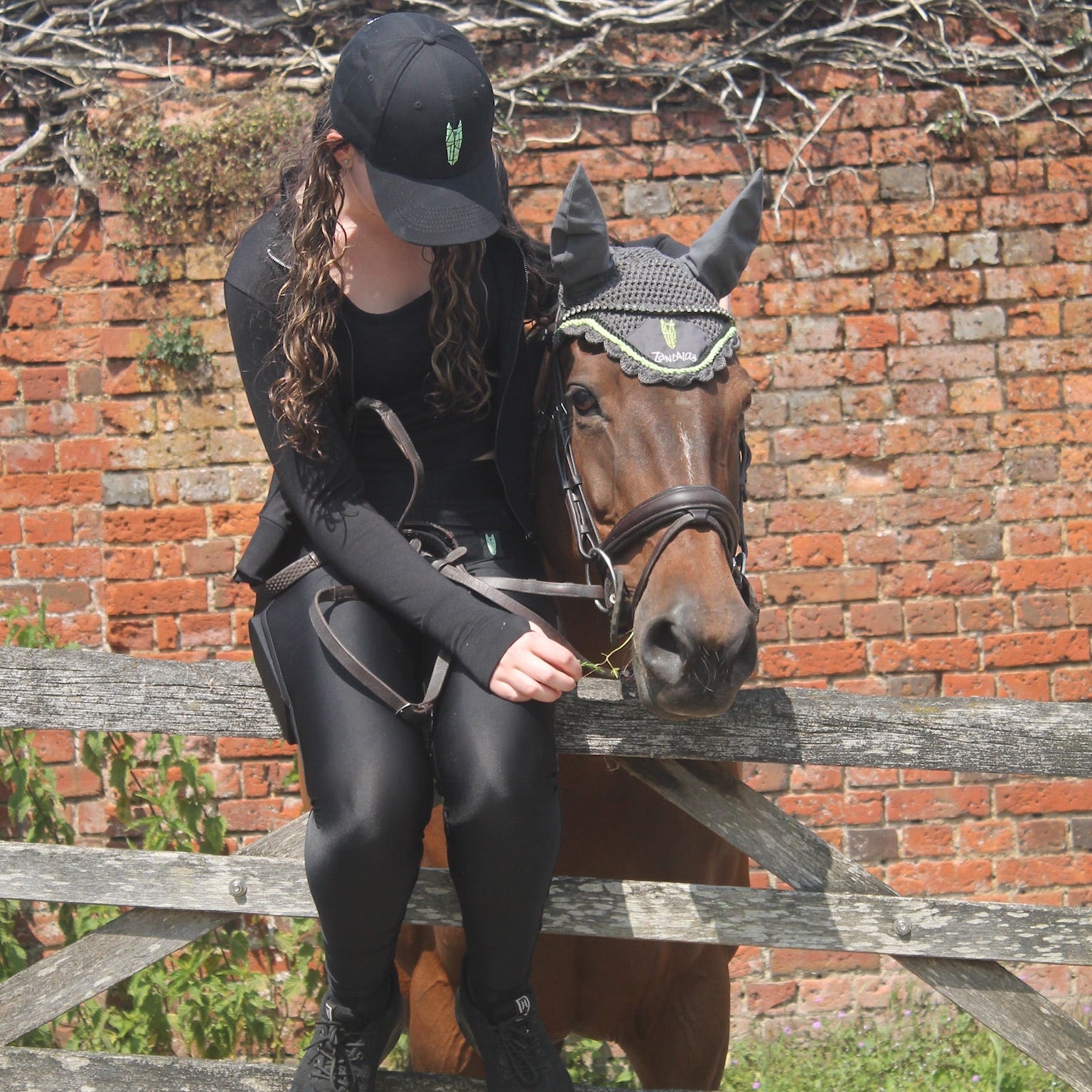 Teenage girl wearing equestrian gear standing next to a horse with a bridle, against a brick wall.