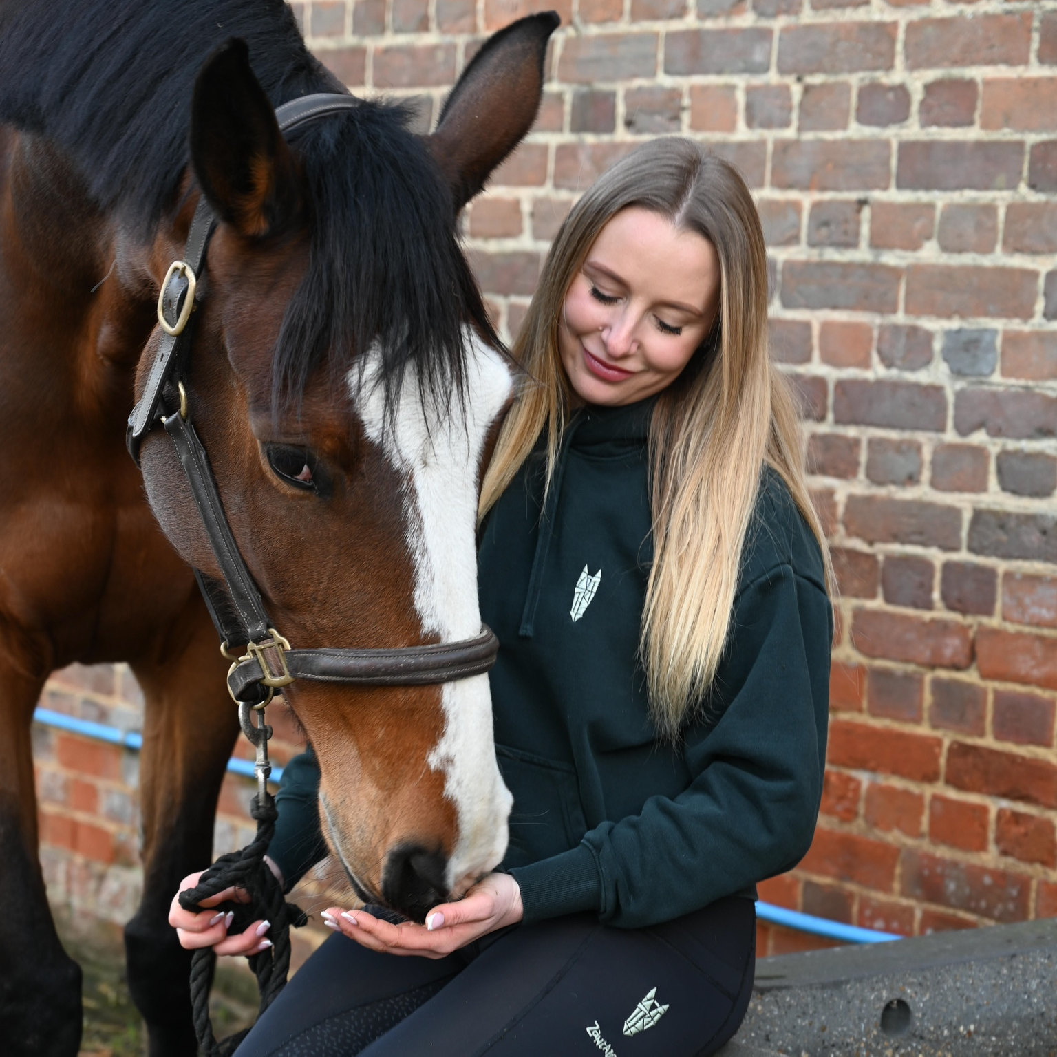 Woman in equestrian attire, wearing a bottle green hoodie interacting with a horse against a brick wall.