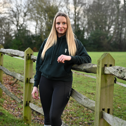 Woman wearing a bottle green hoodie standing by a wooden fence in an outdoor setting