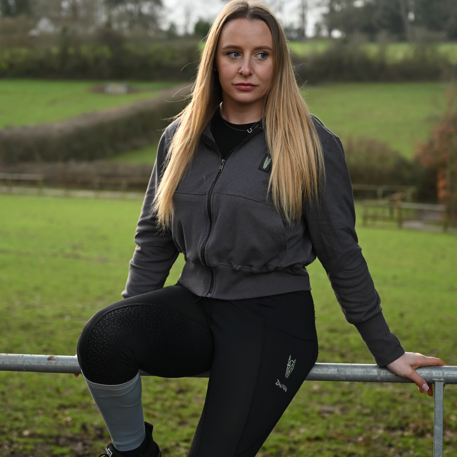 Woman in equestrian wear, wearing a supersoft grey equestrian hoodie posing outdoors on a grassy field