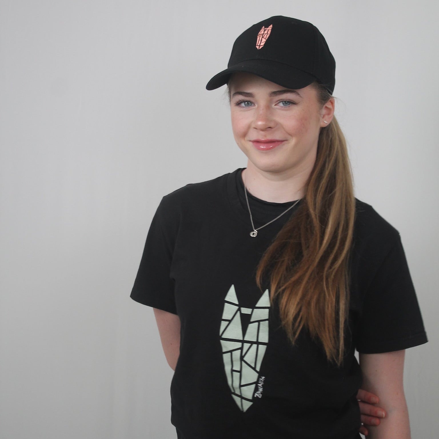 A teenage girl wears an equestrian outfit including a black baseball cap with a dusky pink equestrian logo and a black t-shirt with a large silver green equestrian logo