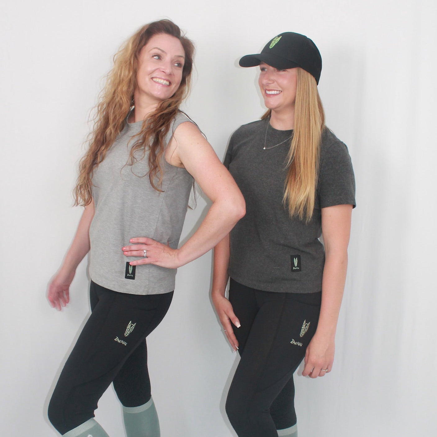 Two women pose in grey tshirts, one in a light grey sleeveless tshirt with black riding leggings and the other in a dark grey short sleeved tshirt with black riding leggings