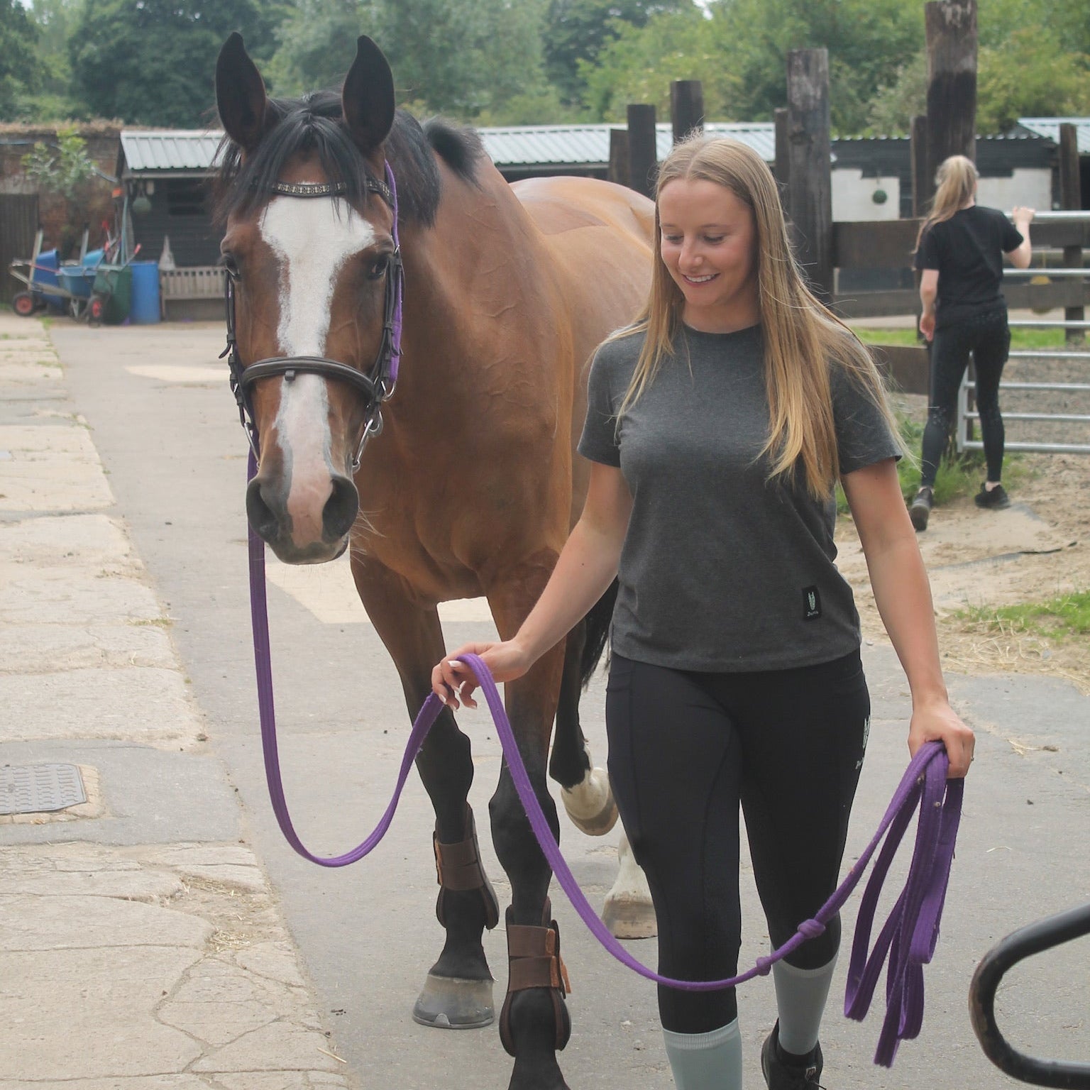 A woman leads a horse on a lunge rope wearing a dark grey t-shirt with a striking grey green equestrian logo