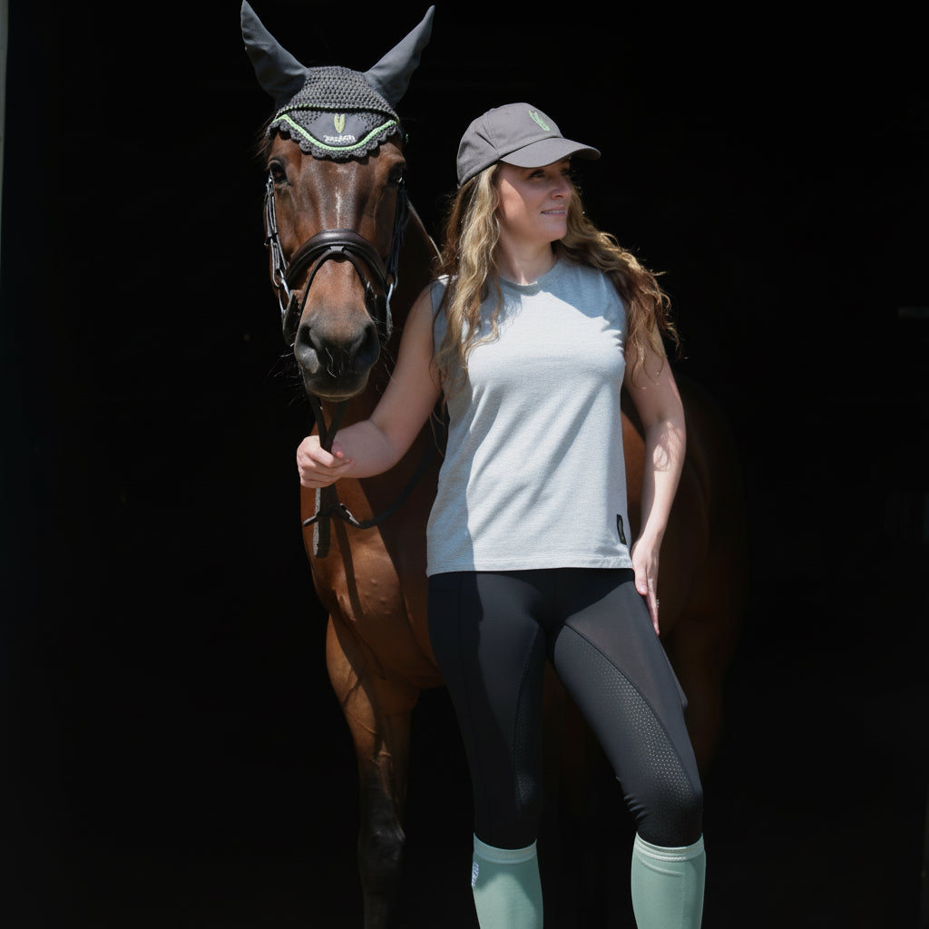 Woman holding a horse in the doorway of a barn wearing a light grey sleeveless tshirt and black riding leggings