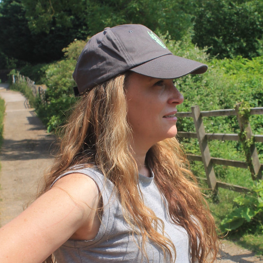 Woman wearing a grey equestrian cap with a silver green horse head logo walking on a dirt path in a natural setting with trees and a wooden fence.