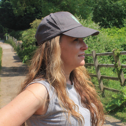 Woman wearing a grey equestrian cap with a silver green horse head logo walking on a dirt path in a natural setting with trees and a wooden fence.