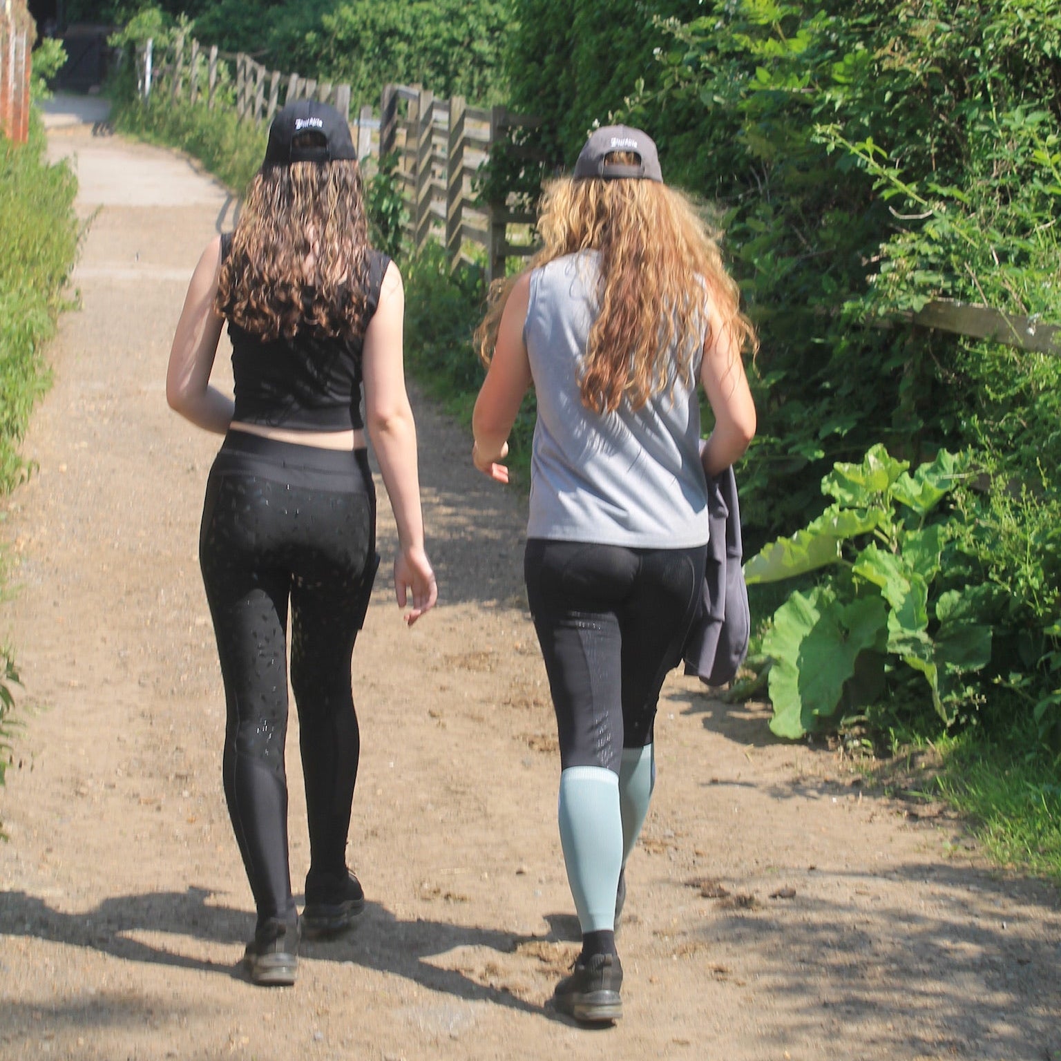 2 women walk down a farm path next to gated fields wearing equestrian clothing, including a sleeveless grey tshirt and riding leggings