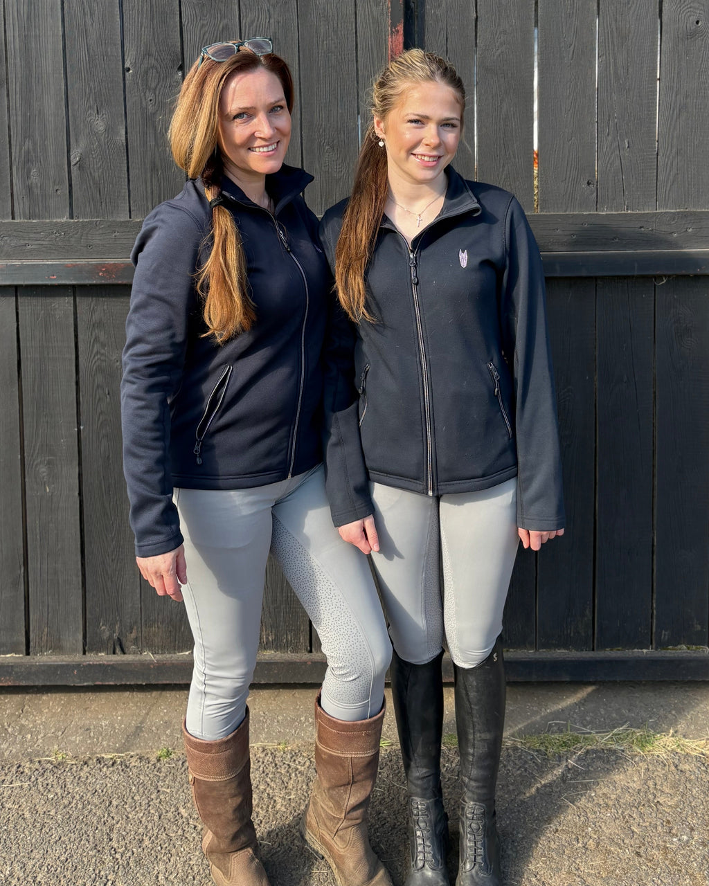 Two women wearing equestrian clothing featuring a geometric horse head logo standing in front of a dark wooden fence
