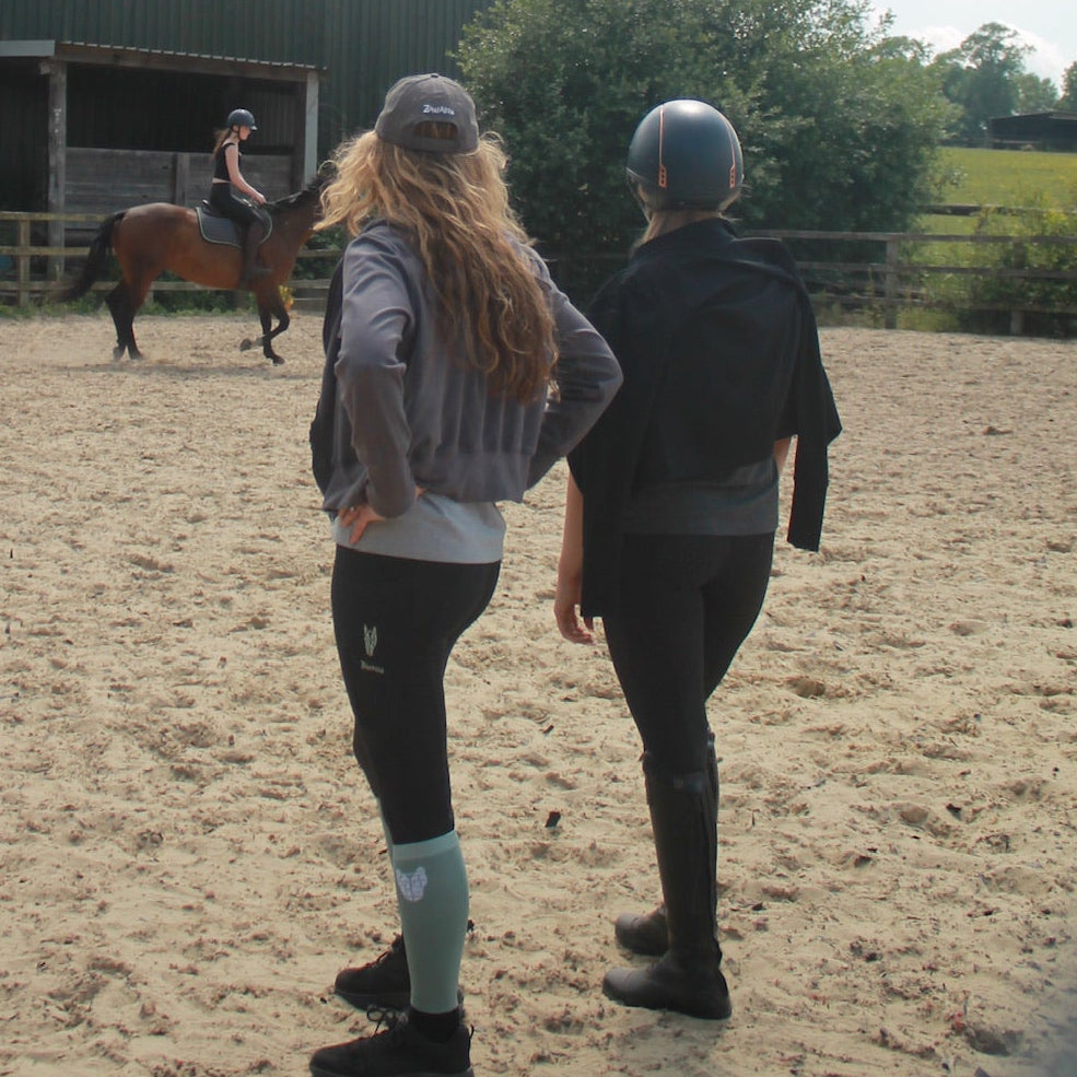 Two women watch a teenager ride a horse wearing an equestrian stretch crop hoodie in grey