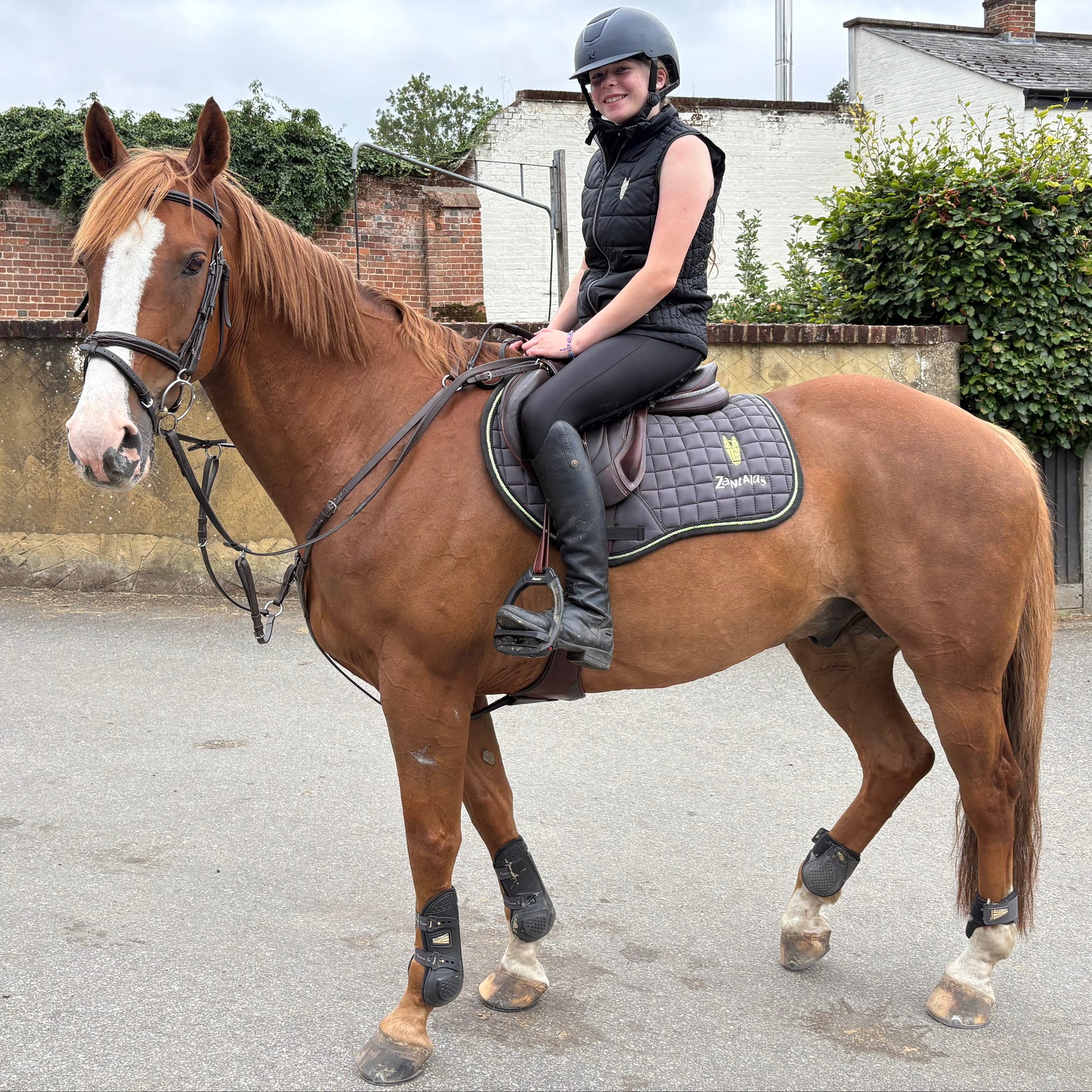 Woman riding a chestnut horse wearing a black eqeustrian gilet bodywarmer on a road with buildings and greenery in the background