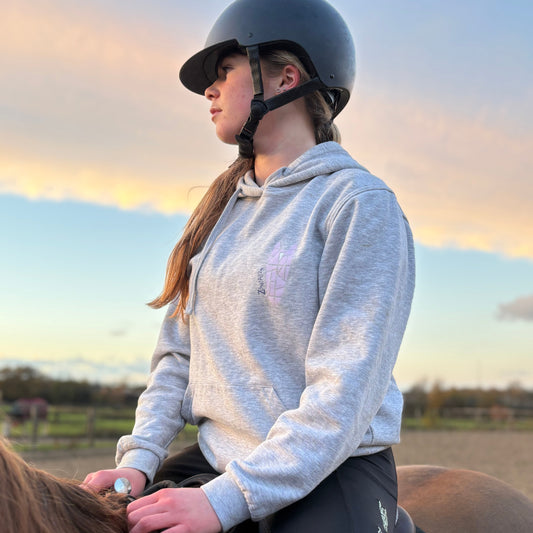 Young woman wearing a light grey hoodie riding a horse in an outdoor setting with a sunset in the background