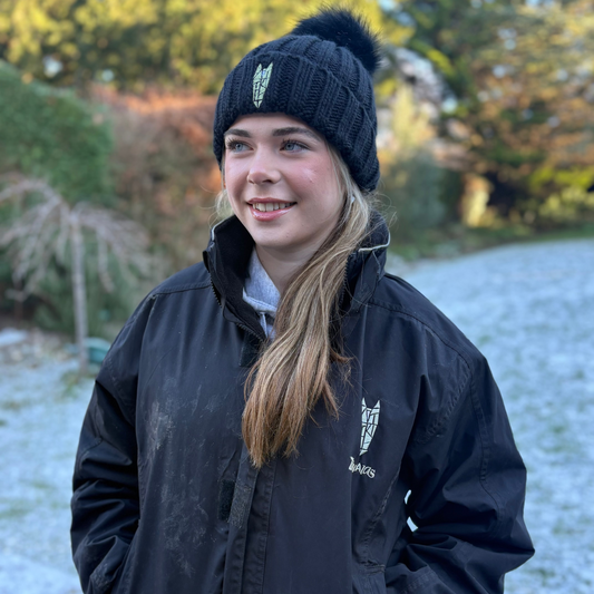 Person wearing a black equestrian jacket and beanie standing outdoors with snow on the ground.