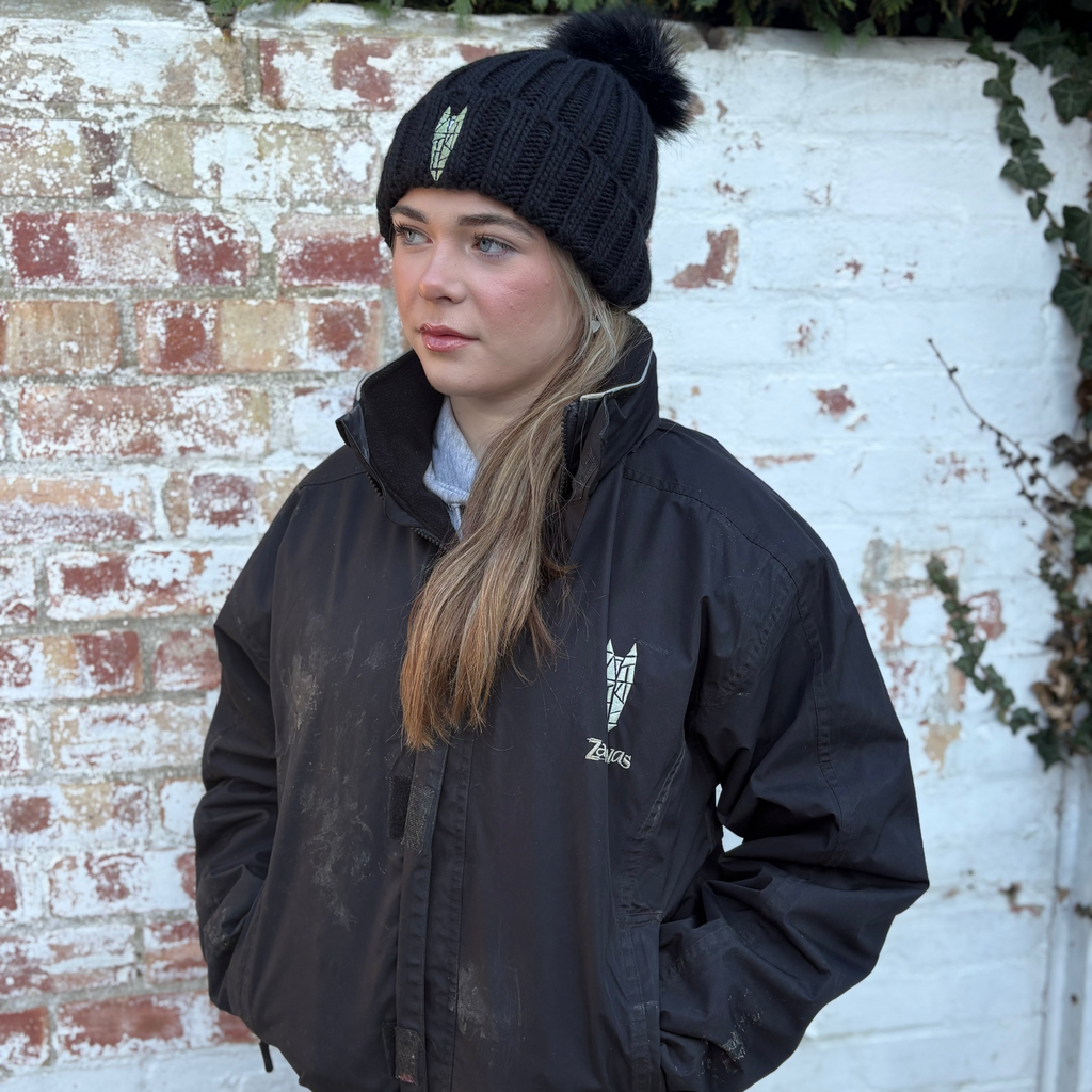 Person wearing a black equestrian jacket and beanie standing against a white brick wall.