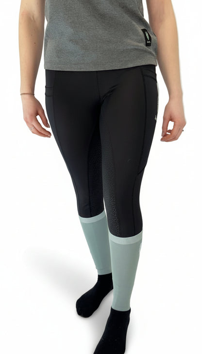 Woman wearing black equestrian riding leggings with a full gel seat and double pockets, view from the front with a striking horse head logo just showing on the left pocket