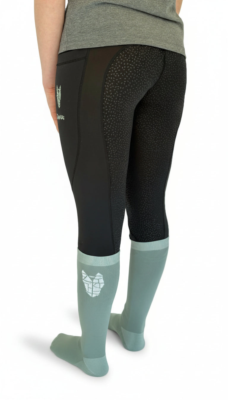 View from the back of a woman wearing black equestrian riding leggings with full gel seat, double pockets and a striking horse head logo on the left pocket