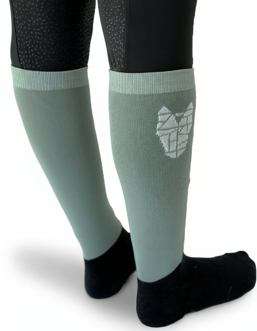 A close up on a woman's calves, wearing black and gray/green equestrian riding socks with a striking white horse head logo on the side