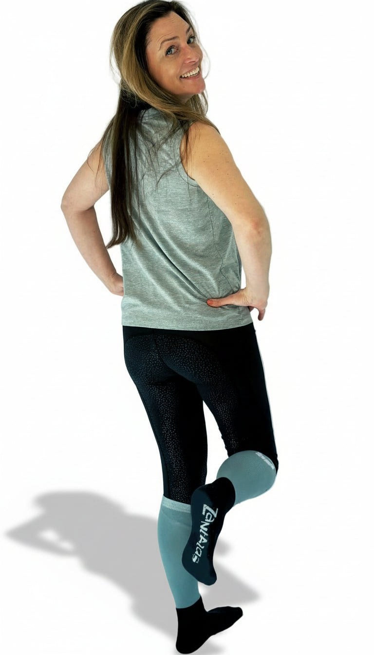 a woman wearing black and gray/green equestrian socks with one foot lifted to show a logo on the bottom of the foot